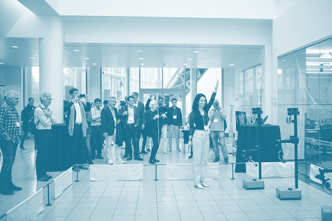 A woman stands in a pen with a group of robots, gesturing to them with her left arm as they dance.